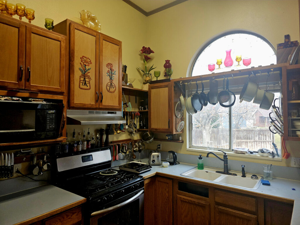 One corner of a very well-organized kitchen. Pots and pans hang on rails over the sink, and cooking utensils hang next to the stove. The cabinet faces are decorated with floral appliques, and colorful glassware is displayed on top.