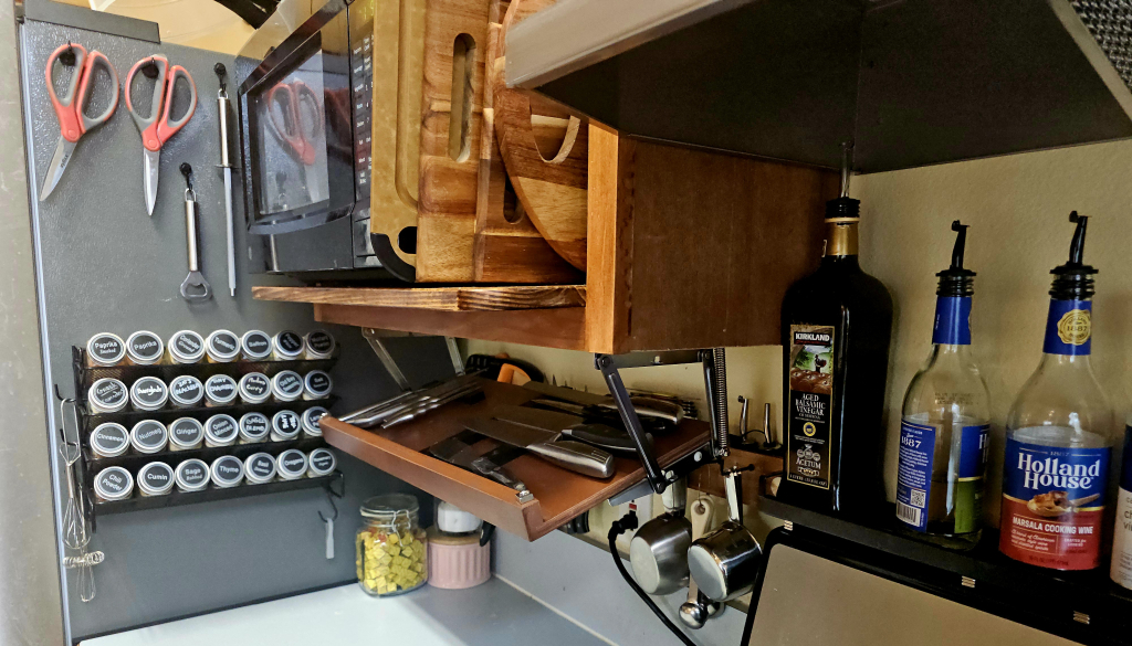 Another corner of the kitchen. A spice rack is magnetically mounted to the side of the refrigerator, with 28 uniformly labeled spice bottles. A drop-shelf is partially lowered from its position beneath the shelf supporting the microwave. The drop-shelf is for knives, all magnetically-docked where they can be hidden and secured from youngsters.